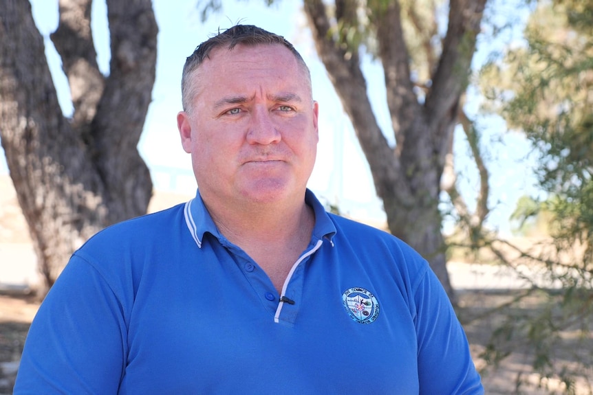 Shane wears a blue shirt while pictured in front of a tree