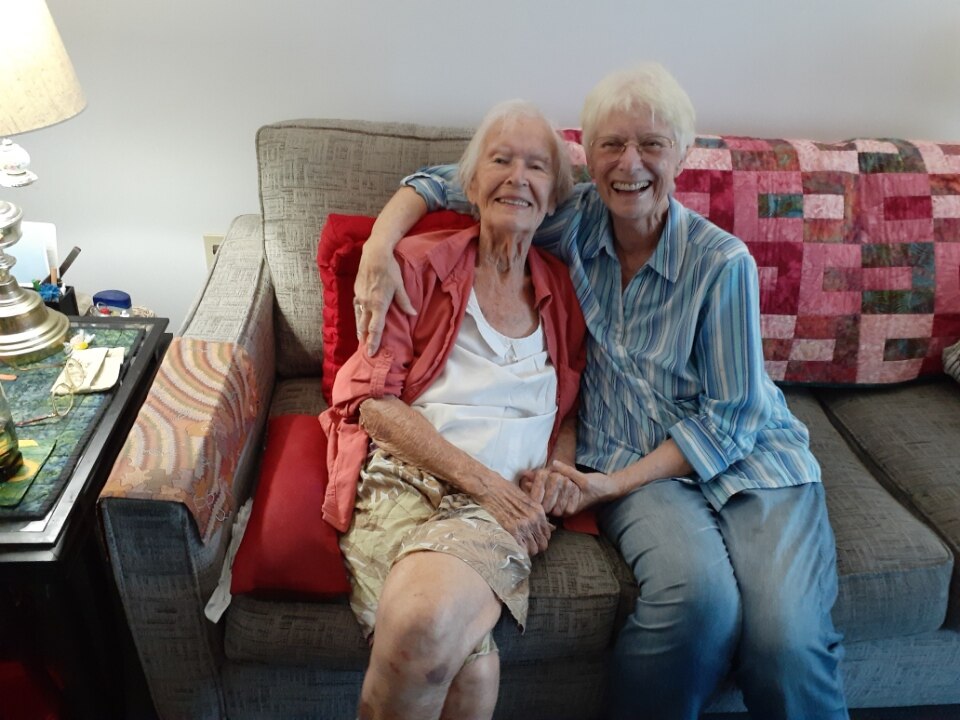 Two women with white hair sit on sofa, holding hands and smiling