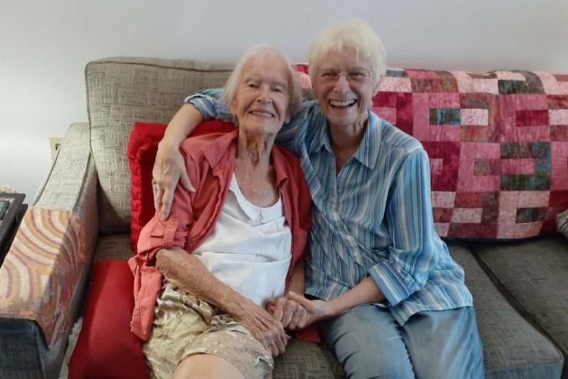 Two women with white hair sit on sofa, holding hands and smiling