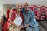 Two elderly women sit smiling and holding hands on a couch.