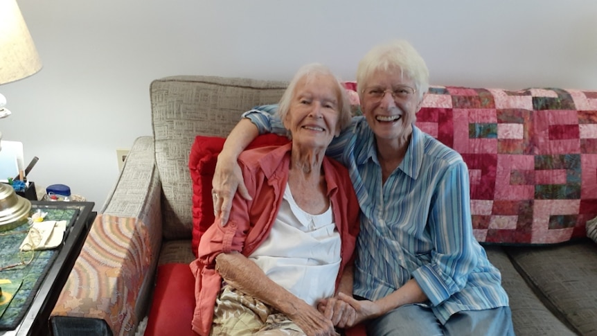 Two elderly women sit smiling and holding hands on a couch.