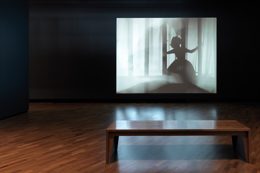 In an empty gallery, a white screen with the blacks silhouettes of a woman amongst trees