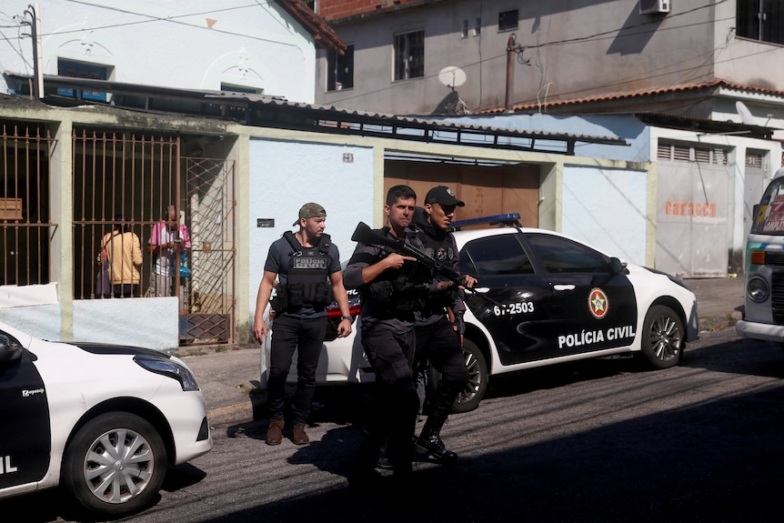 Trois hommes portant un gilet pare-balles et un portant une arme à feu traversent une rue avec des voitures de police en arrière-plan