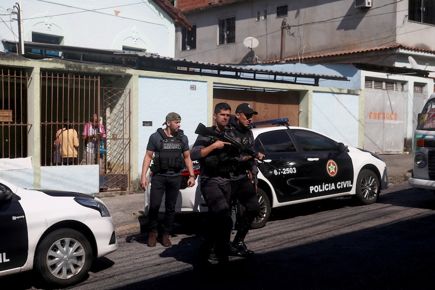 Three men wearing body armour and one carrying a gun cross a street with police cars in the background