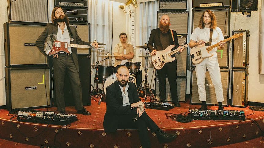 The band IDLES holding their instruments on a small stage in an old pub or club, all wearing suits