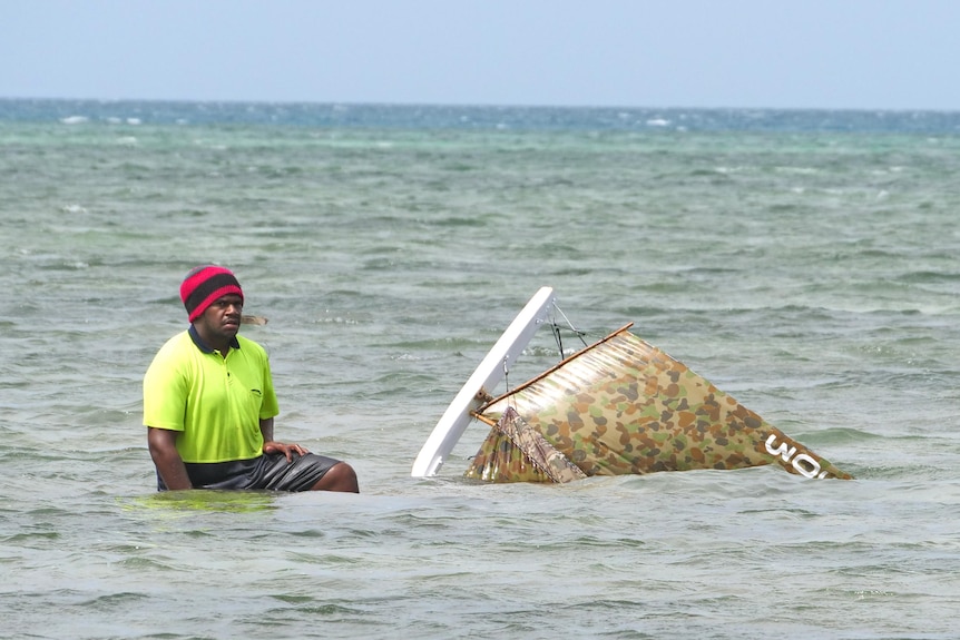 Man kneels in shallow with a boat laying on its side.
