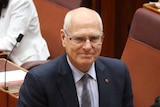 Jim Molan, with his hands crossed in front of him, smiles in the Senate. Marise Payne is sitting behind him.