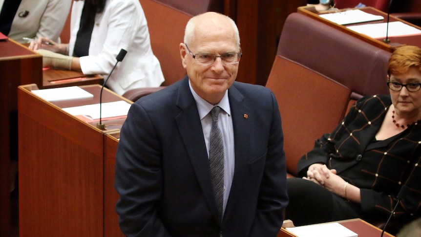 Jim Molan, with his hands crossed in front of him, smiles in the Senate. Marise Payne is sitting behind him.
