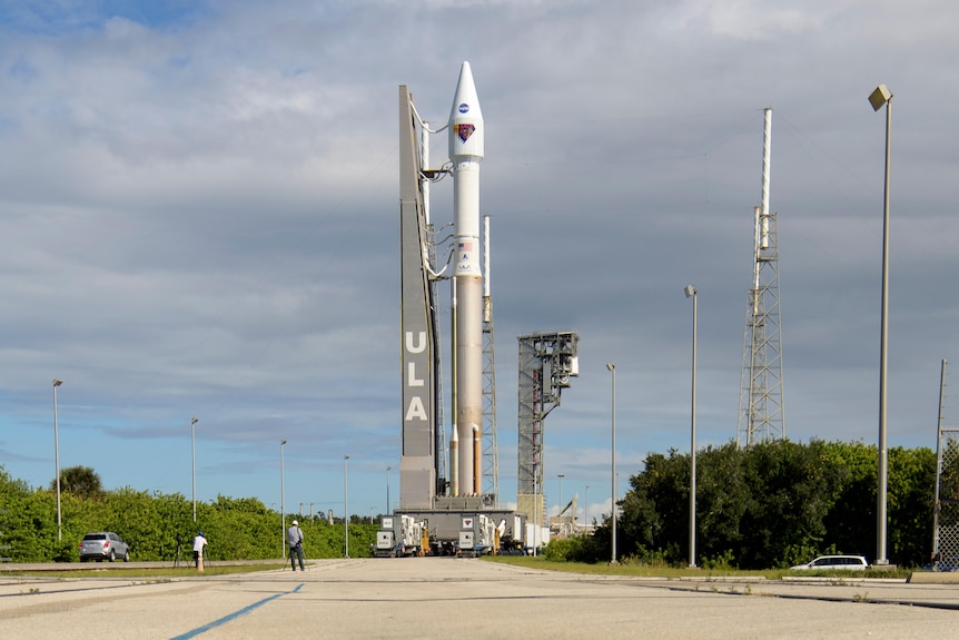 The Atlas V rocket stands ready to launch, its length reflected in a puddle in the foreground