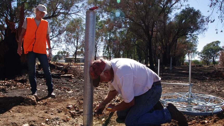 A man in a white t-shirt is on his knees wiring a new fence post