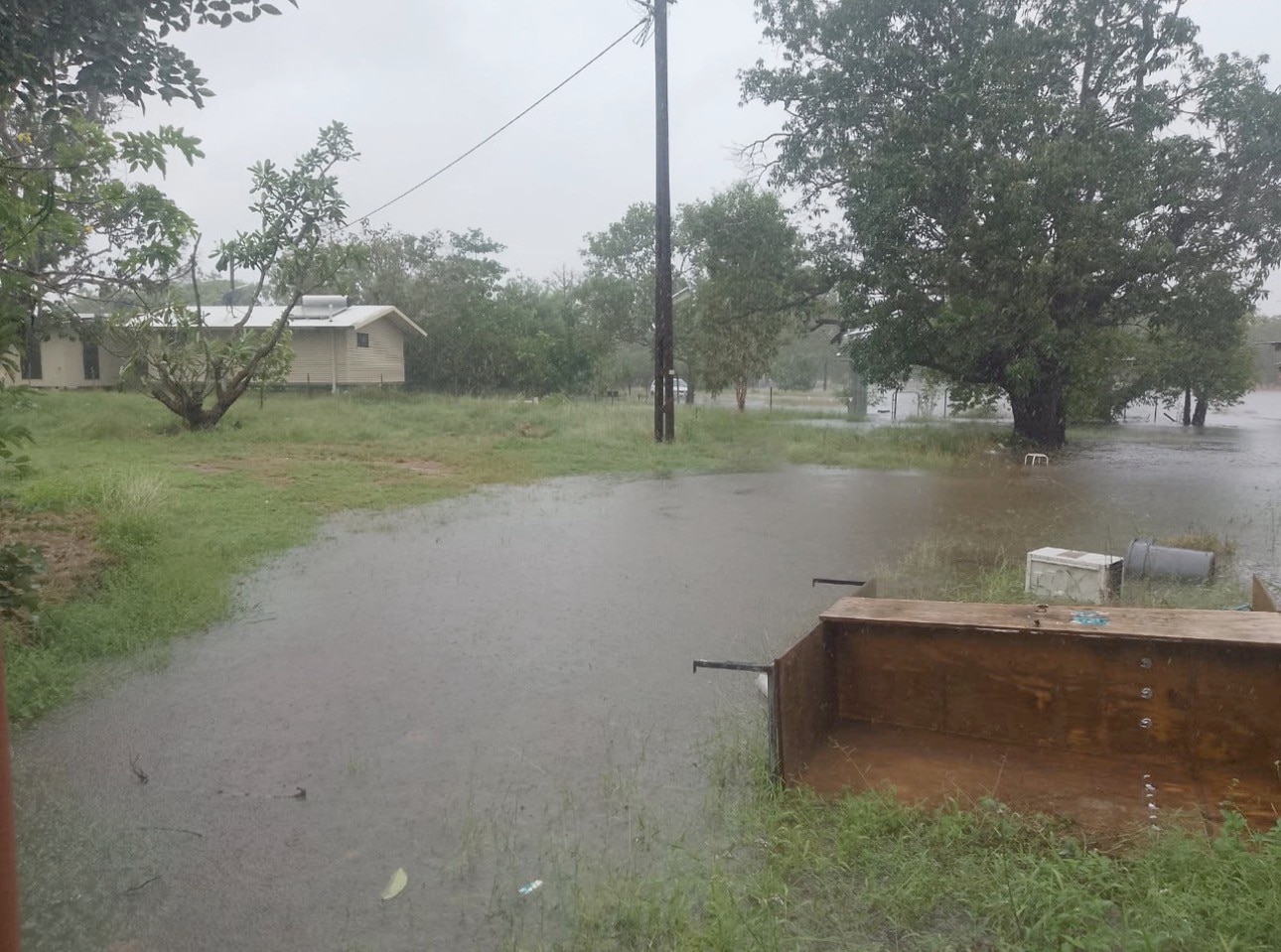 Flood waters winding live a river around trees, with housing to left of image, gray sky, gray flood waters.