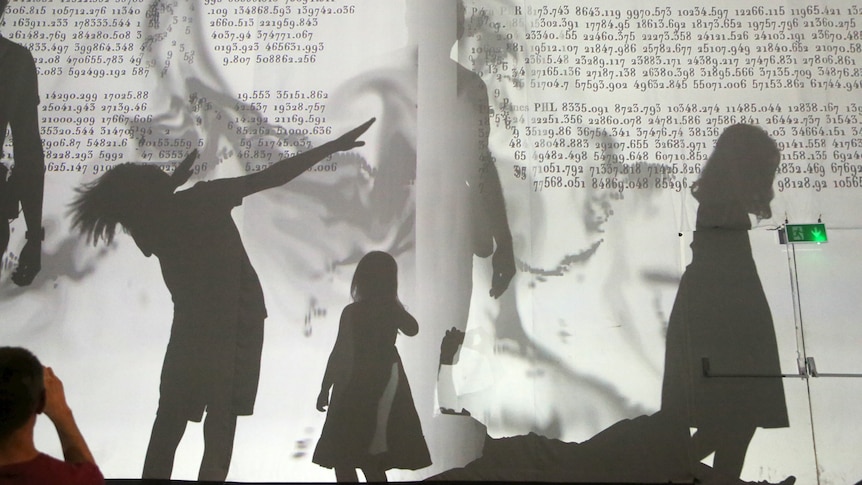 Three children's shadows are cast onto a wall lit up with the projection of a 19th century book.