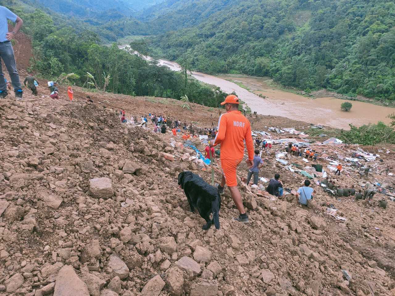 A man with a black dog walking over rubble with multiple other people in the background.