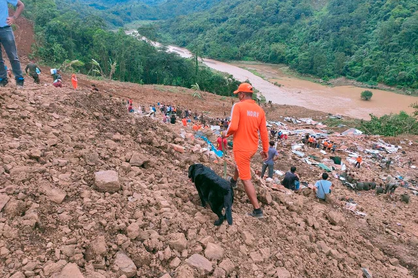 A man with a black dog walking over rubble with multiple other people in the background.