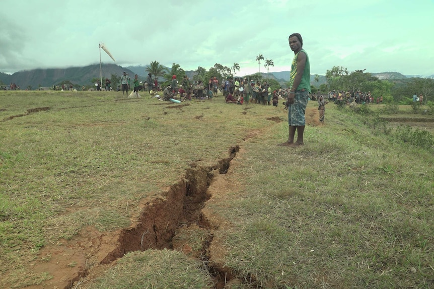 A man stands next to a crack in the earth, with several other cracks visible in the background