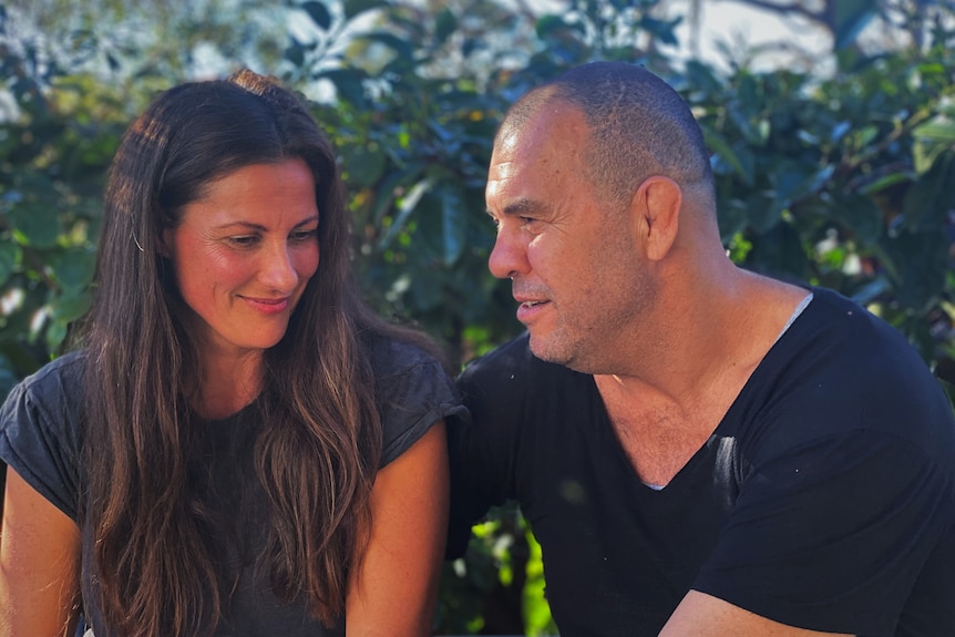 A dog, woman and man sit together on an outdoor couch, smiling.