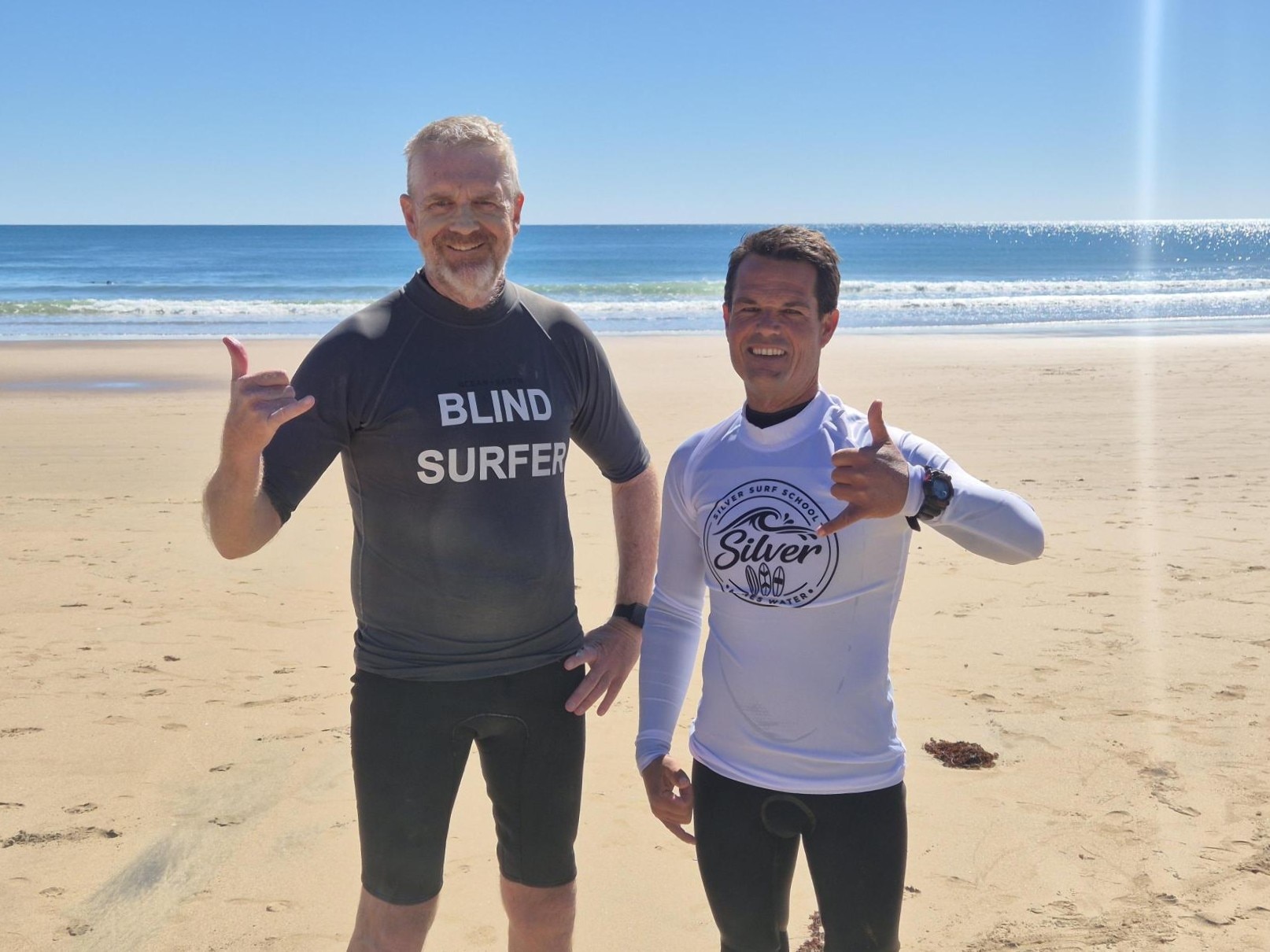 Two male surfers standing side by side holding up the shakka hands with the ocean in the background
