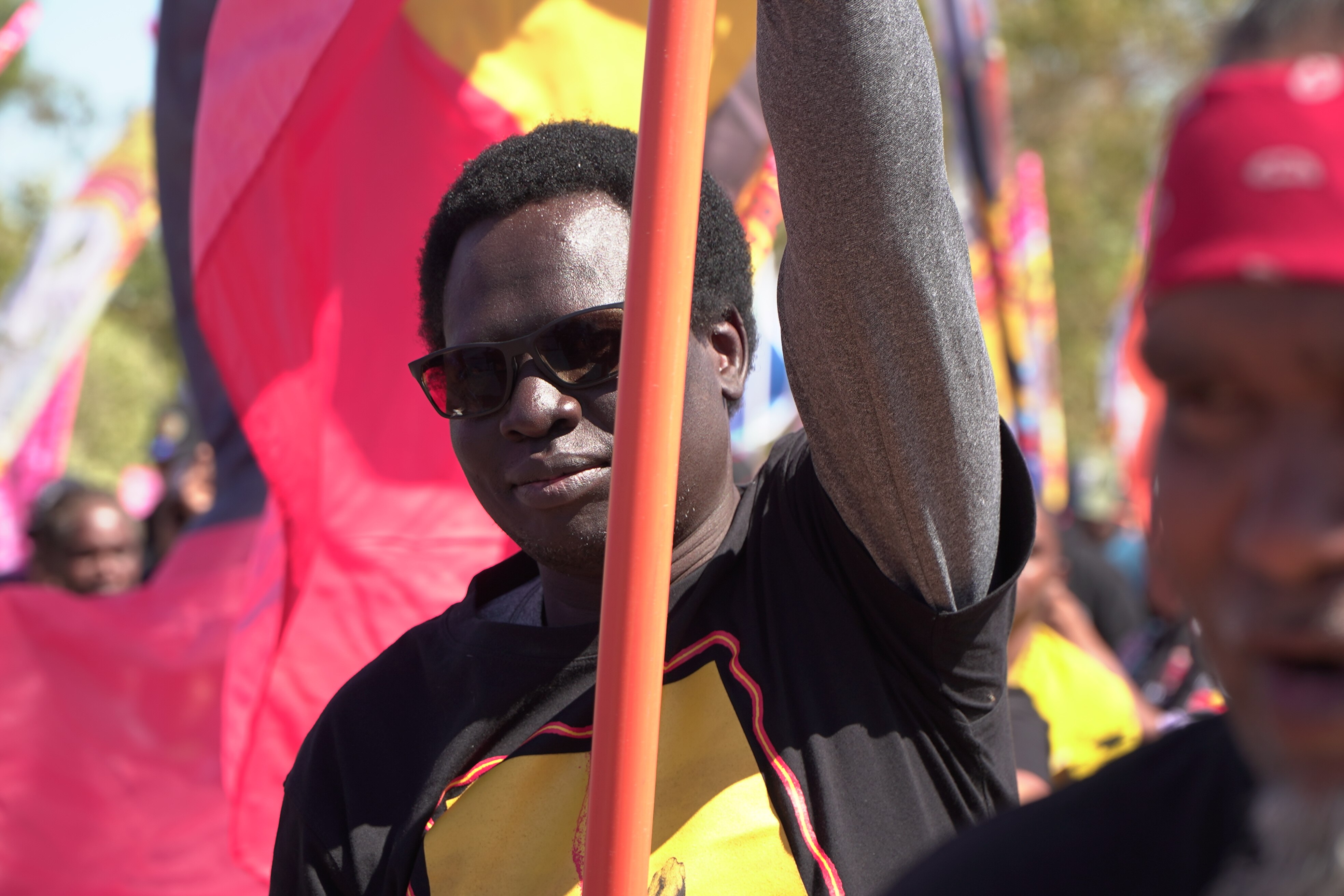 A man with dark complexion, black short tight curls, black sunglasses, wearing black shirt and holding pole for Aboriginal flag.