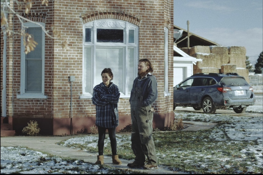 A woman in plaid shirt and boots stands next to a man in overalls outside a house, snow on the ground