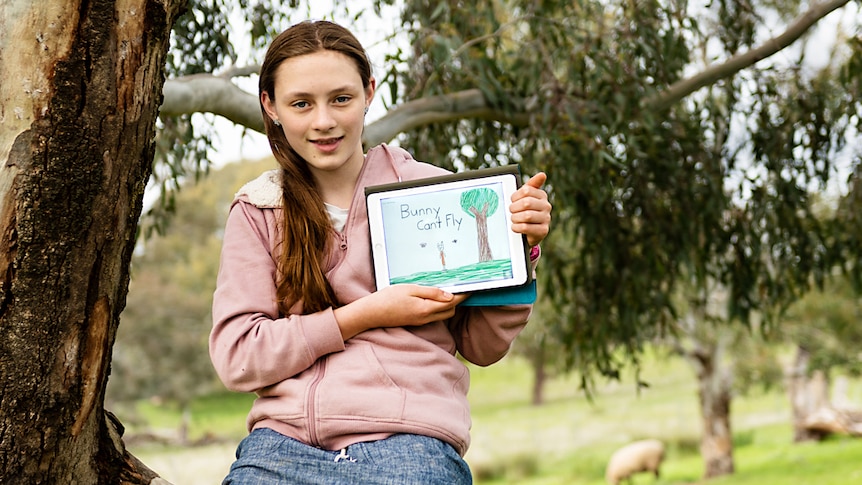 A girl sits in a tree holding an tablet displaying the cover of an eBook whose title says Bunny Can't Fly.