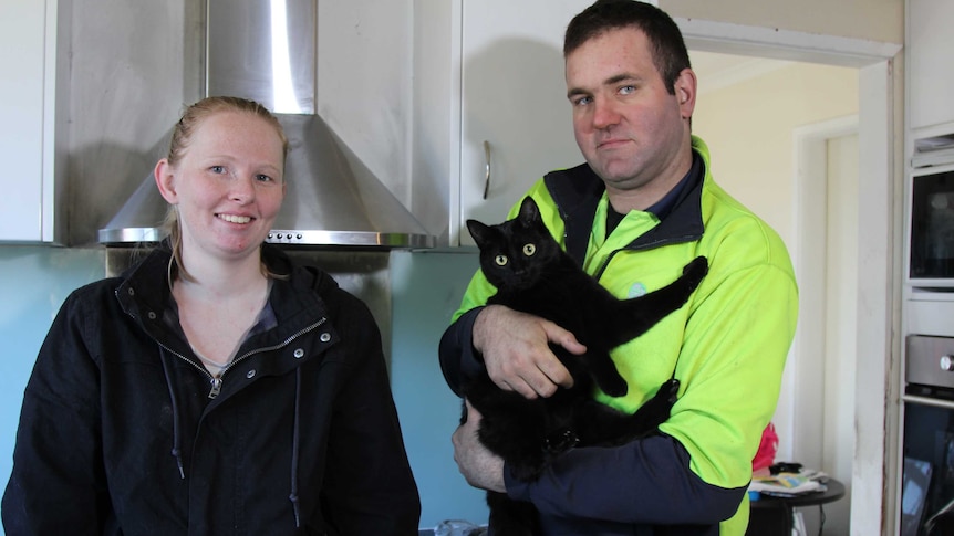 A couple with their pet cat. The man looks annoyed as he holds the cat.