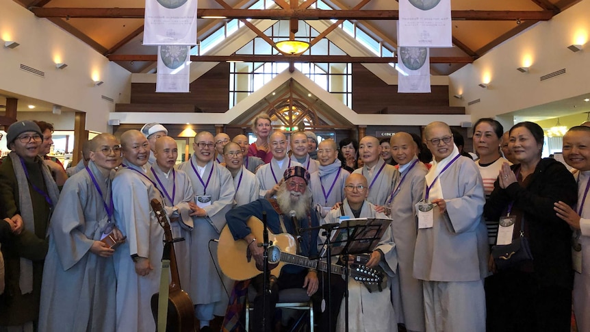 Korean nuns dressed in light coloured robes. A man with a guitar and white beard sits in the centre.