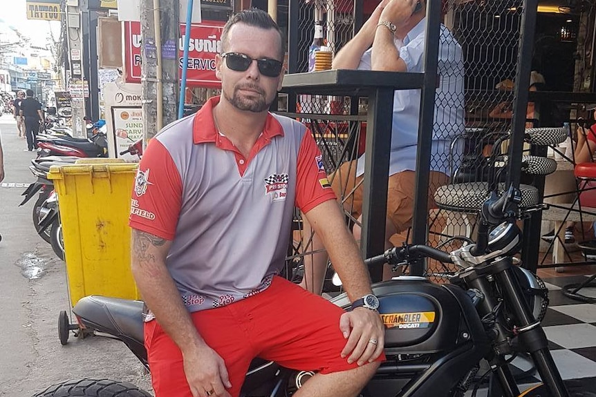 Luke Joshua Cook sitting on a motorbike on the side of a Thai road wearing red shorts and a grey shirt.