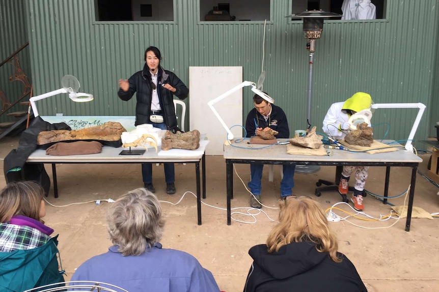 Three people working on dinosaur bones in front of a small audience.