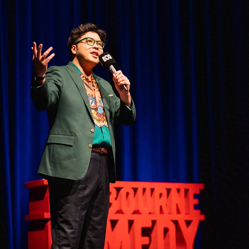 Kuah Jenhan on stage with a red Comedy Festival sign and dark blue stage curtains in the background.