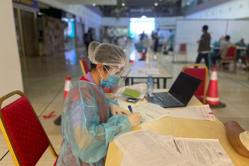 A Laotian healthcare worker in personal protection equipment sits waiting for people to register for vaccinations
