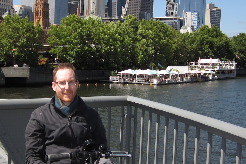 A man sits on a three-wheeled scooter next to a river with trees and city buildings in the background