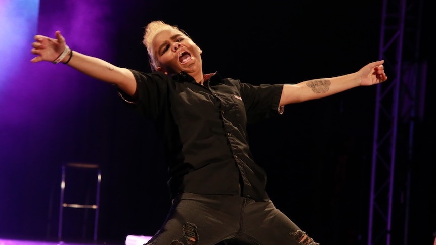 A drag king sliding on his knees with his arms wide on stage