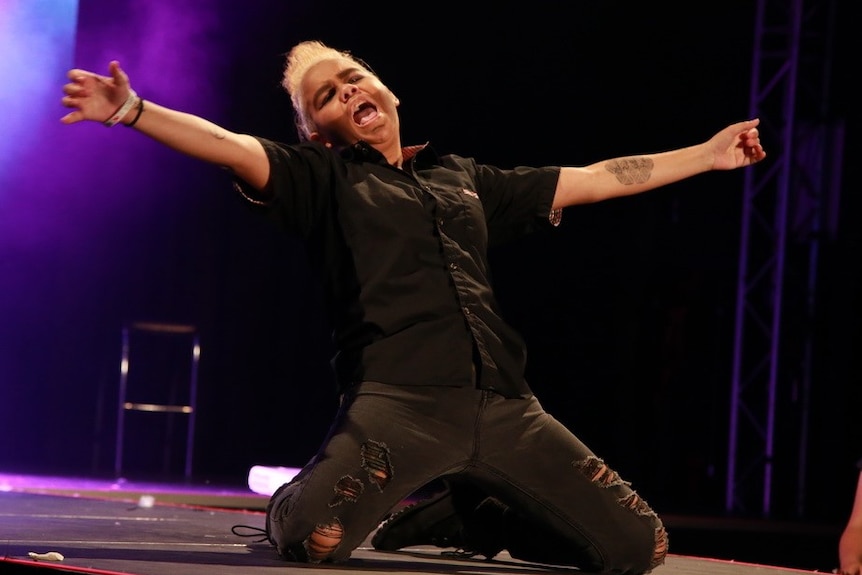 A drag king sliding on his knees with his arms wide on stage