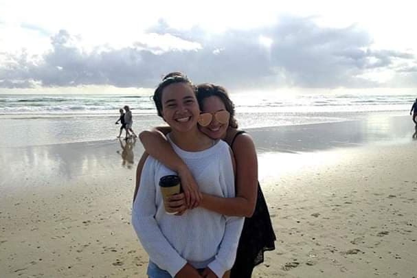 A smiling woman in sunglasses hugs her teenage daughter from behind at a sunny beach.