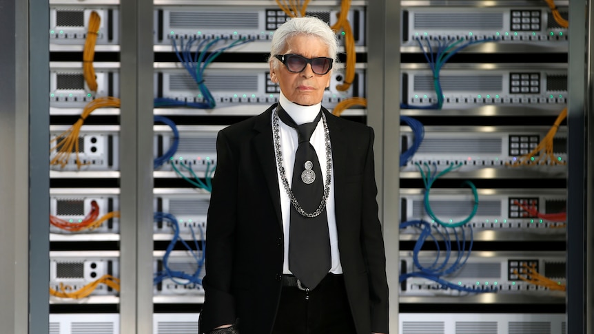 Karl Lagerfeld standing in front of a technological backdrop, wearing a black suit, dark sunglasses and fingerless gloves