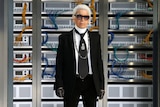 Karl Lagerfeld standing in front of a technological backdrop, wearing a black suit, dark sunglasses and fingerless gloves