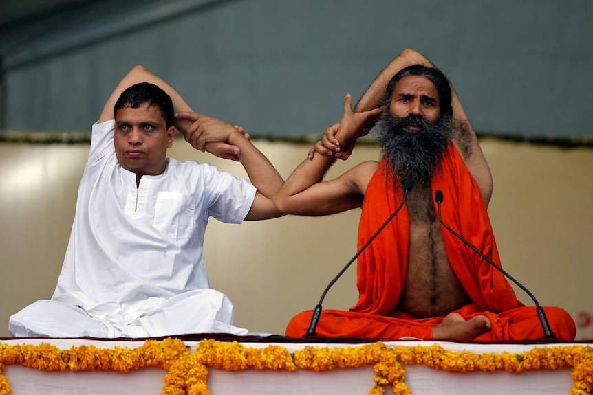 Two middle-aged Indian men sit cross-legged in a yoga pose.