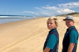 Two boys aged 11 standing on an empty beach looking toward the camera with a long stretch of beach behind them, flags in distanc