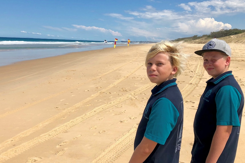 Two boys aged 11 standing on an empty beach looking toward the camera with a long stretch of beach behind them, flags in distanc