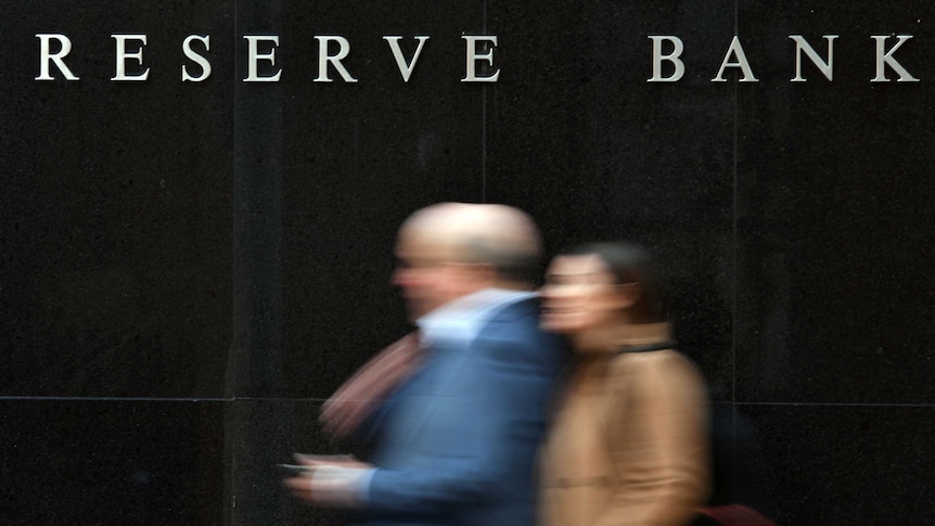 Two blurred people walking past a black wall with 'Reserve Bank' in white lettering.