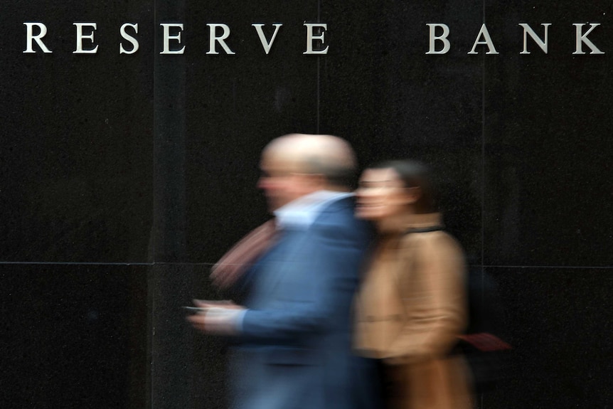Two blurred people walking past a black wall with 'Reserve Bank' in white lettering.