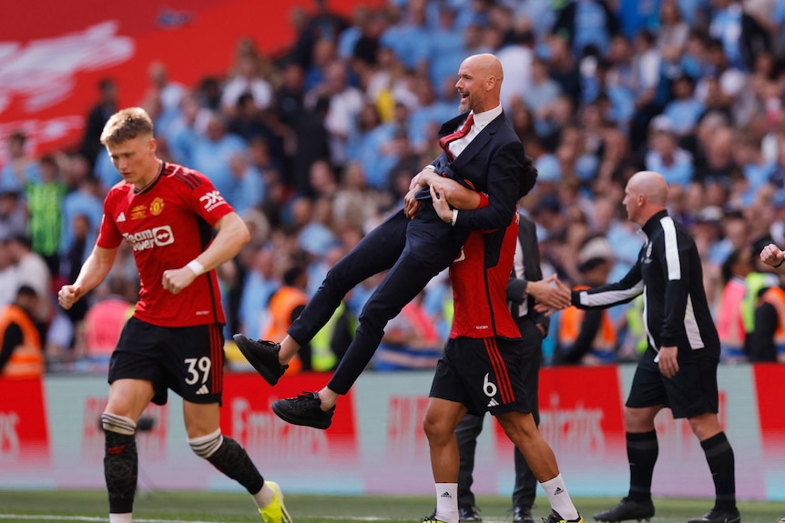 A football manager wearing a suit is picked up by a player in celebration.