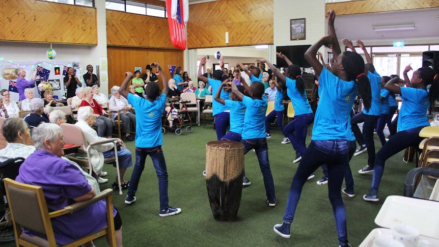 African children's choir sing and dance before a crowd of elderly people.
