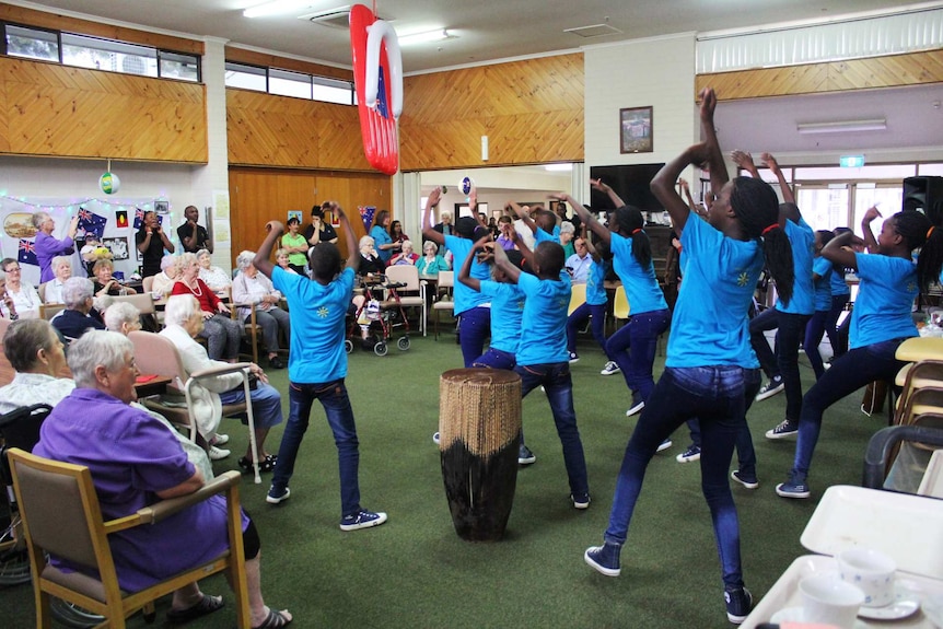 African children's choir sing and dance before a crowd of elderly people.