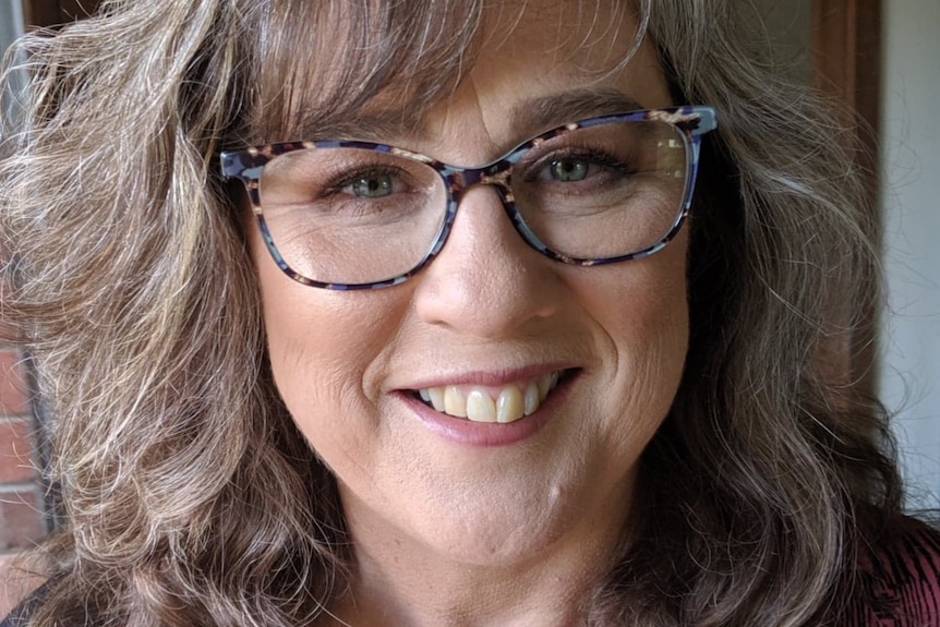 A woman smiles, she has fluffy curled brown hair, blue glasses and mauve lipstick. 
