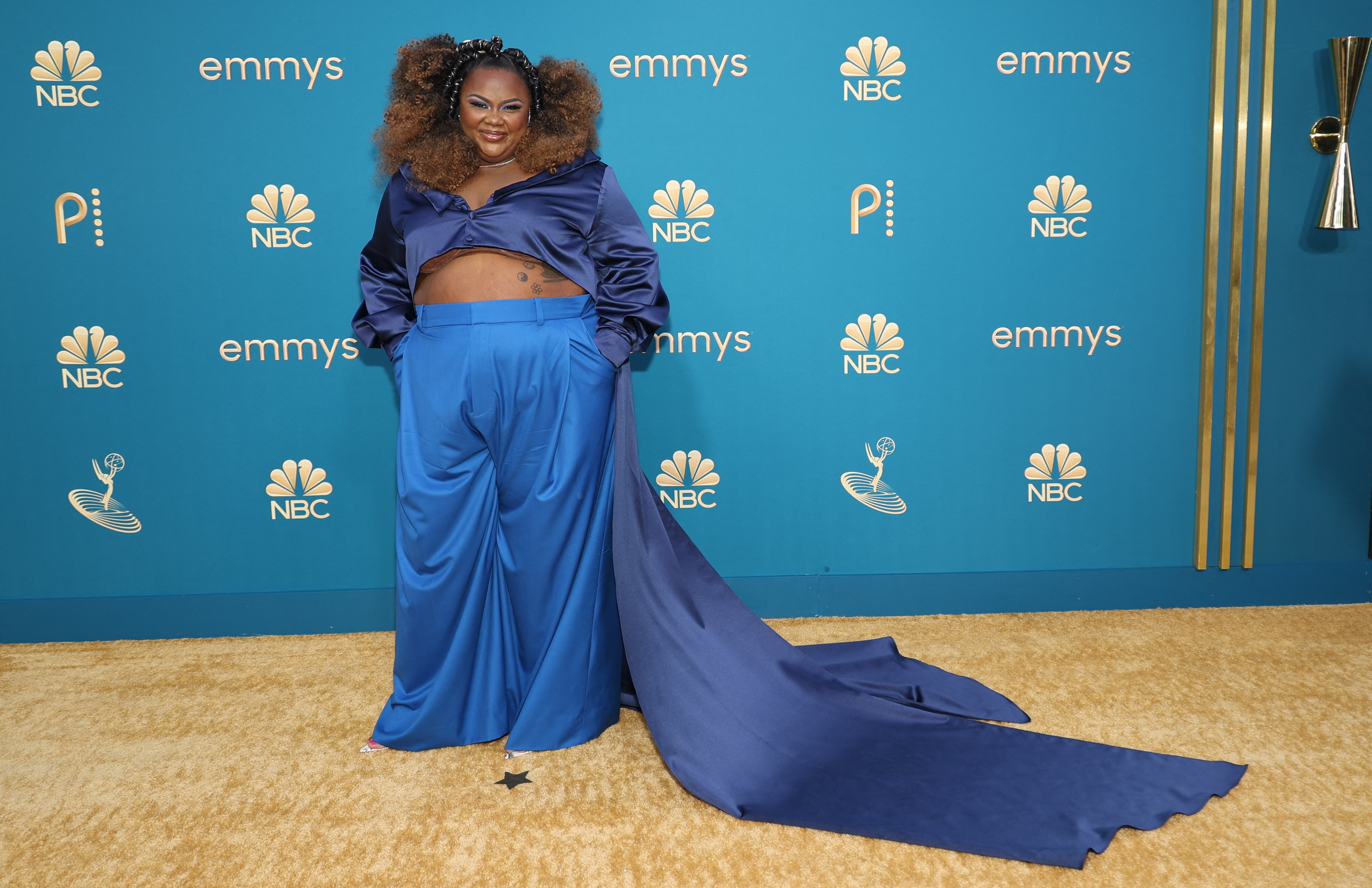 Nicole Byer wearing a pair of blue silky trousers and a dark blue top with two long trains. 