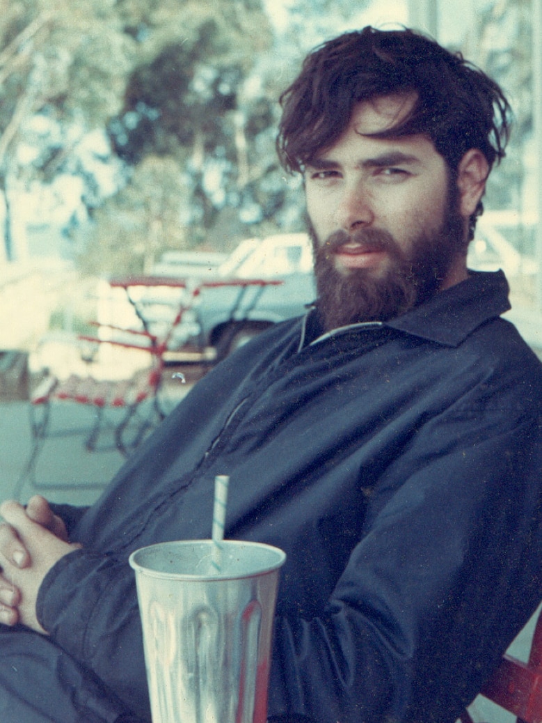 A young man with a beard looks seriously at the camera, a tin milkshake cup at his arm.