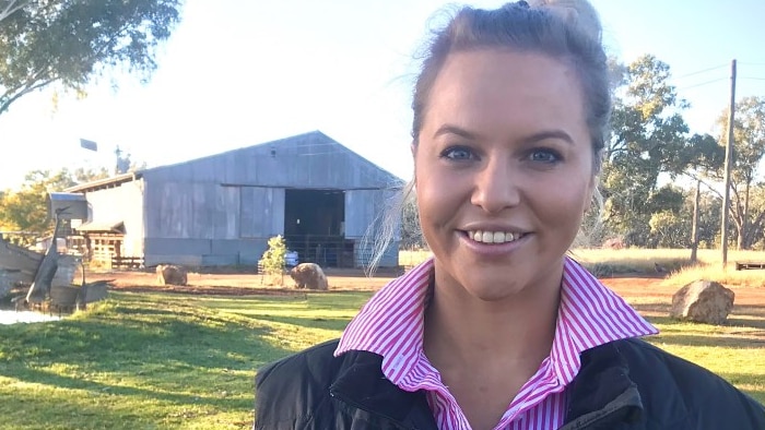 A woman of with dark hair smiles for the camera. She stands near a shed on a farm.