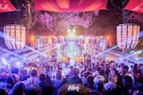 A crowd faces a colourful stage underneath diagonal strobe lights and colourful banners.