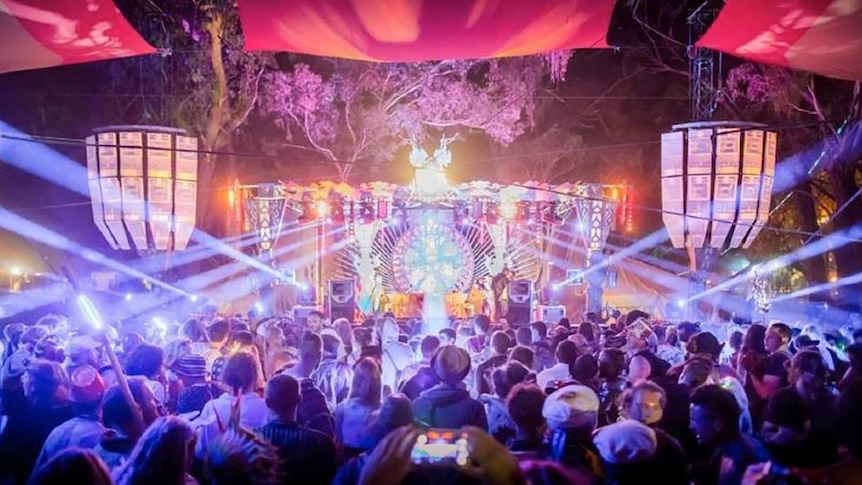 A crowd faces a colourful stage underneath diagonal strobe lights and colourful banners.